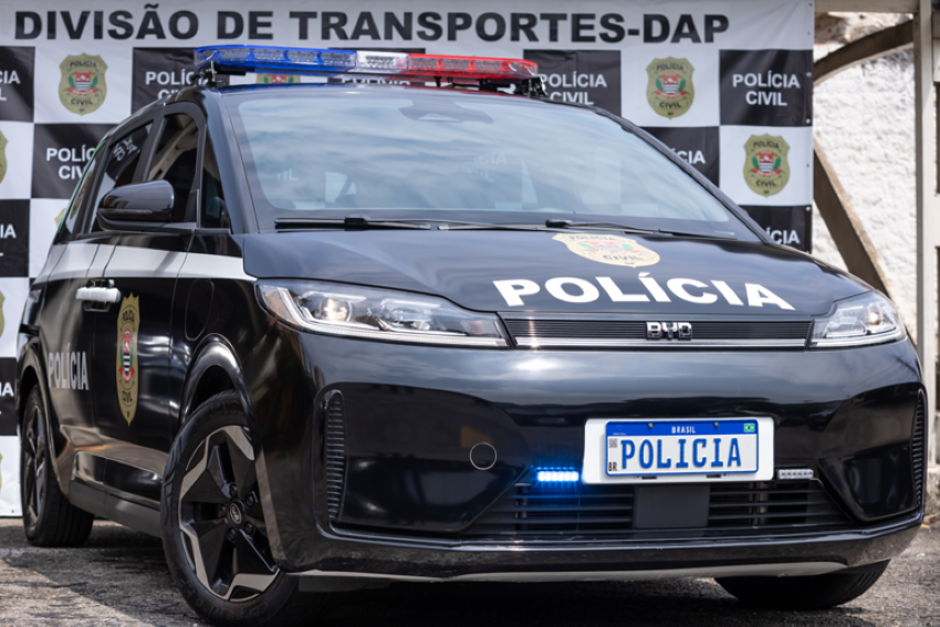 Carro elétrico da BYD com os adesivos da polícia na frente de um painel com vários símbolos da polícia civil. Na placa do carro se lê "polícia"