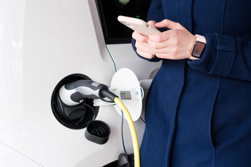 close de um carro elétrico sendo carregado, ao lado uma pessoa com roupa social e relógio digita mexe no celular.