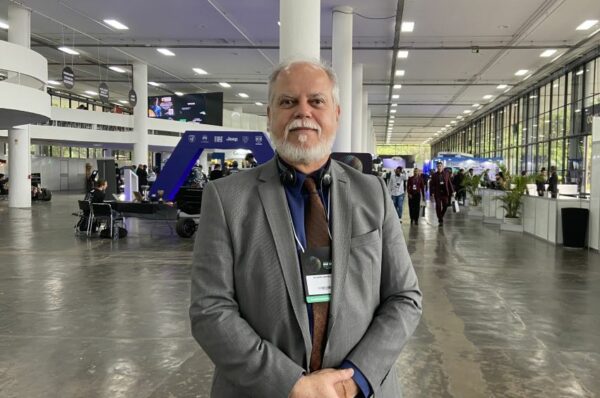 Ricardo Bacellar é um homem de cabelos brancos e barba branca, e está vestido com um terno cinza em meio a um evento