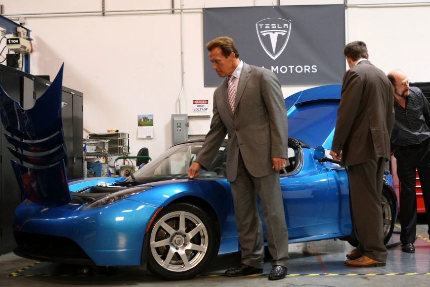 Arnold Schwarzenegger é um homem forte e alto, e na foto está com a mão direita em um carro Tesla