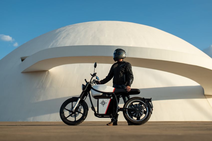 Motociclista vestido de preto segura moto branca da Origem em frente a construção em formato circular
