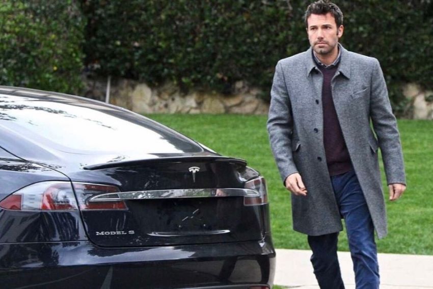 Vestindo um blazer cinza, ator Ben Affleck anda por trás do Tesla Modelo S de cor preta