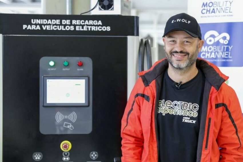 Thiago Castilha, de barba grisalha e boné, posa para foto ao lado de estação de recarga