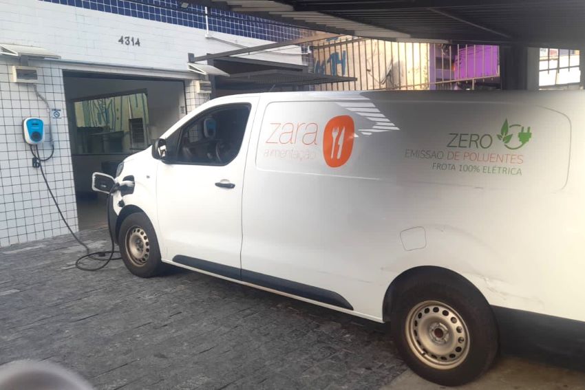 Furgão elétrico branco com o logotipo da Zara Alimentação em laranja na lateral recebe recarga na sede da empresa