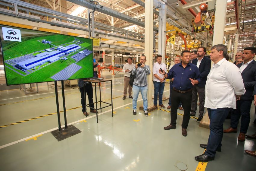 Dentro da fábrica, governador Tarcísio Freitas, de camisa branca, observa televisor com imagens do prédio