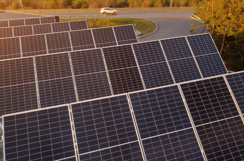 Painéis fotovoltaicos para geração de energia solar estão posicionados ao lado de rodovia, com veículo branco passando ao fundo