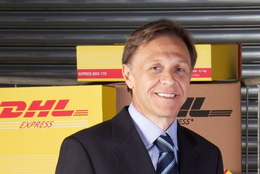Amaury Vitor tem cabelos curtos e usa terno e gravata em frente a caixas da DHL