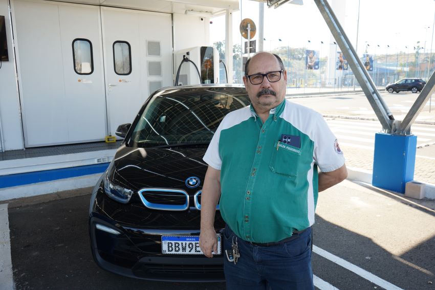 Carlos Figueiredo é um homem calvo, de óculos e bigode, e veste uma camisa verde, à frente de um BMW i3