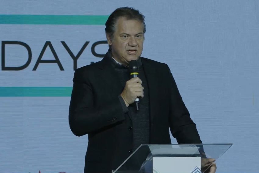Vestido todo de preto, Márcio Lima Leite fala com microfone em palco de evento