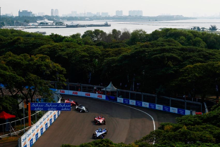 Carros da Fórmula E contornam curva durante e-Prix de Jacarta, em meio a paisagem com árvores e lago ao fundo