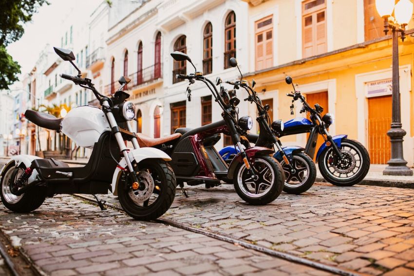Quatro modelos de scooters estão lado a lado em rua de paralelepípedos e prédios coloridos ao fundo