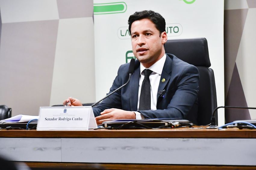 De terno e gravata, Rodrigo Cunha fala em microfone sentado atrás de uma mesa
