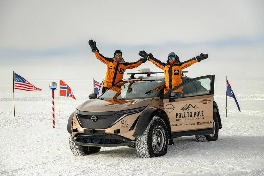 Chris e Julie estão de pé, em cima do Nissan Ariya, ao lado de bandeiras, na estrutura que marca o Polo Sul, na Antártida