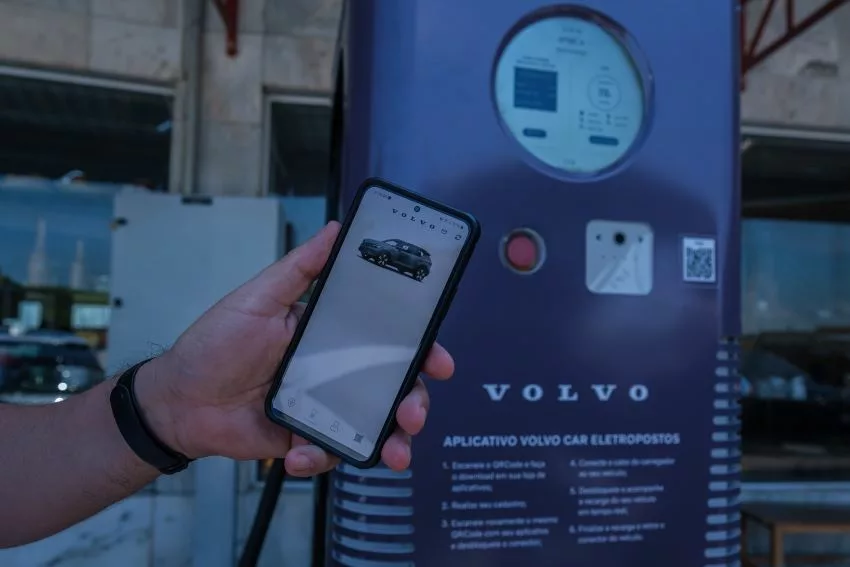 Imagem mostra mão de homem segurando Smartphone conectado no aplicativo da Volvo em frente a um carregador