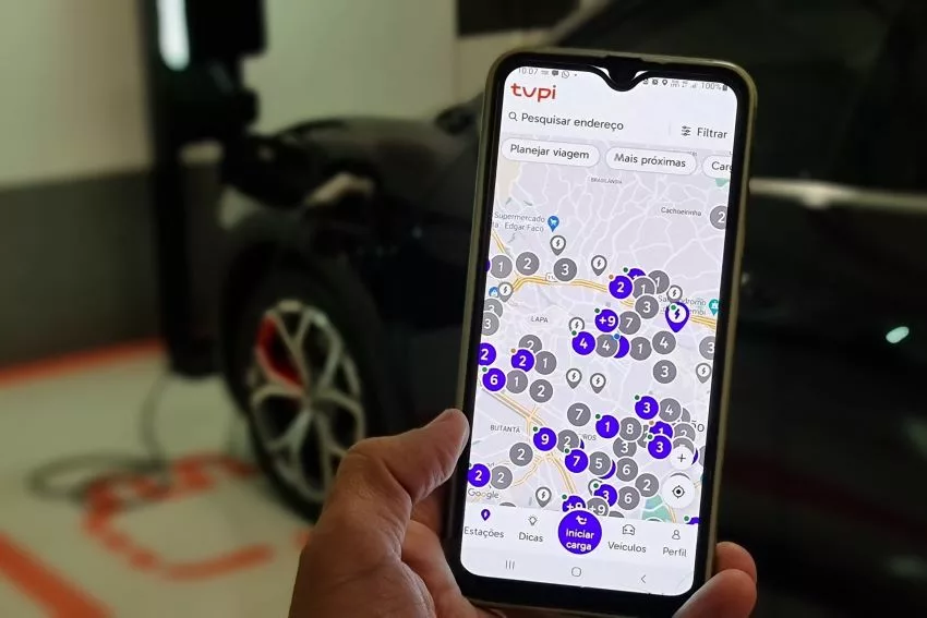 Imagem mostra detalhe do aplicativo Tupi Recarga em smartphone, com carro elétrico desfocado ao fundo