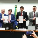 Deputados exibem relatórios atrás de uma mesa e a bandeira do Brasil ao fundo