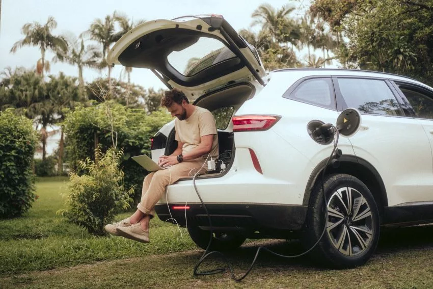 Homem sentado no porta-malas do carro utiliza notebook com energia proveniente do carro