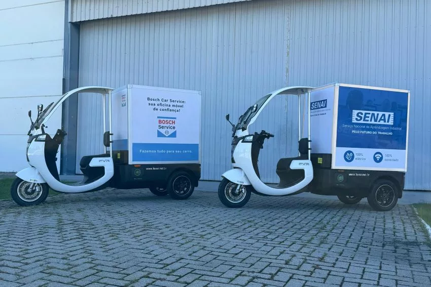 Dois triciclos da Fever com box acoplados estão estacionados em frente a portão de fábrica