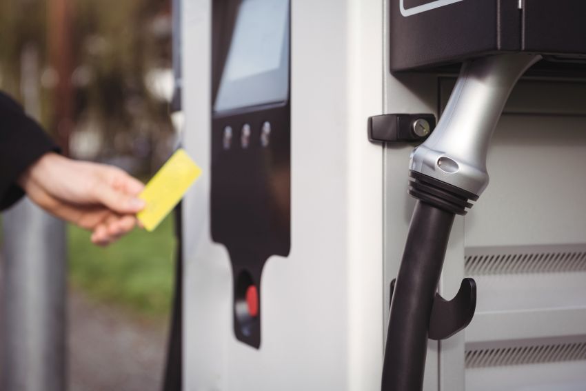 Imagem mostra mão de um homem segurando cartão de crédito próximo a uma estação de recarga de VE