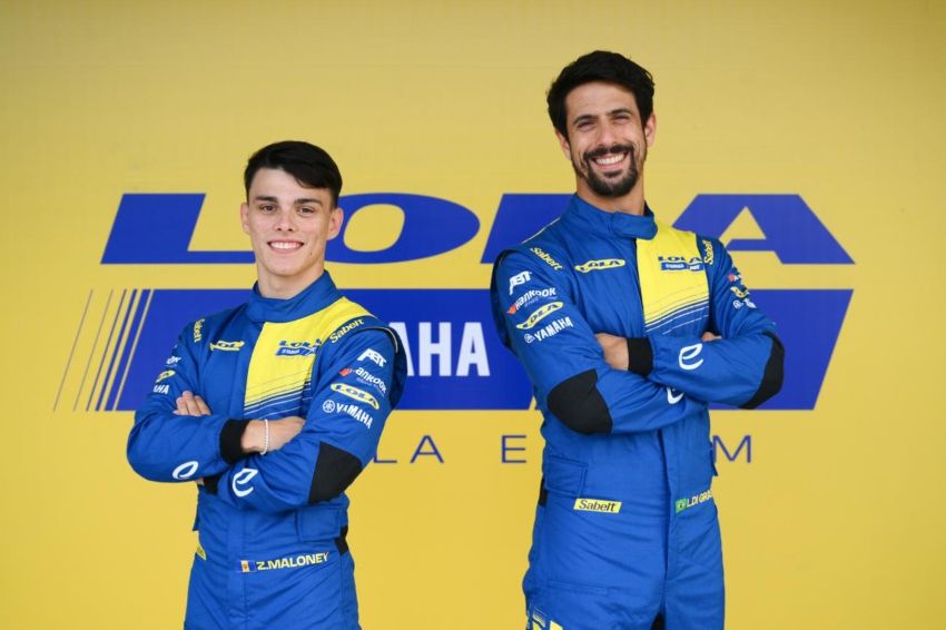Os dois pilotos posam para foto em frente a mural amarelo com a marca da Lola ao fundo